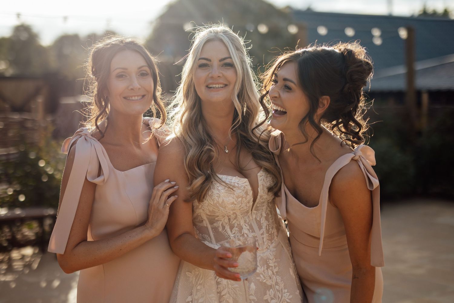 A bride in a lace gown poses joyfully with two bridesmaids in blush dresses in golden sunlight.