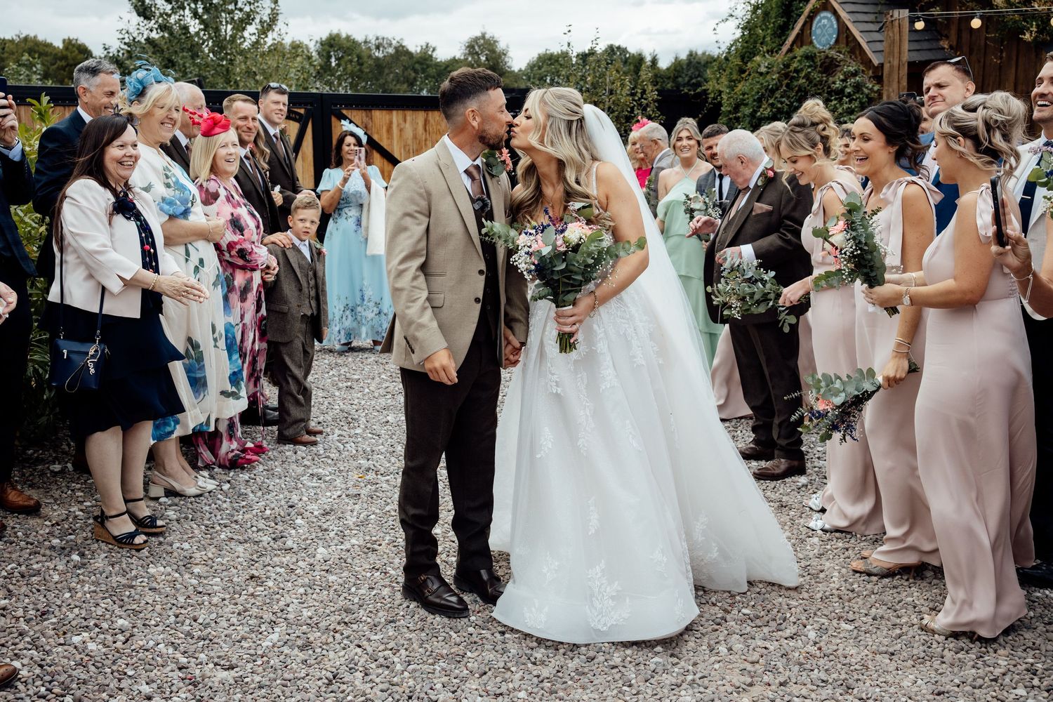 Bride and groom share a kiss on gravel path lined with cheering wedding guests and bridesmaids in blush pink.