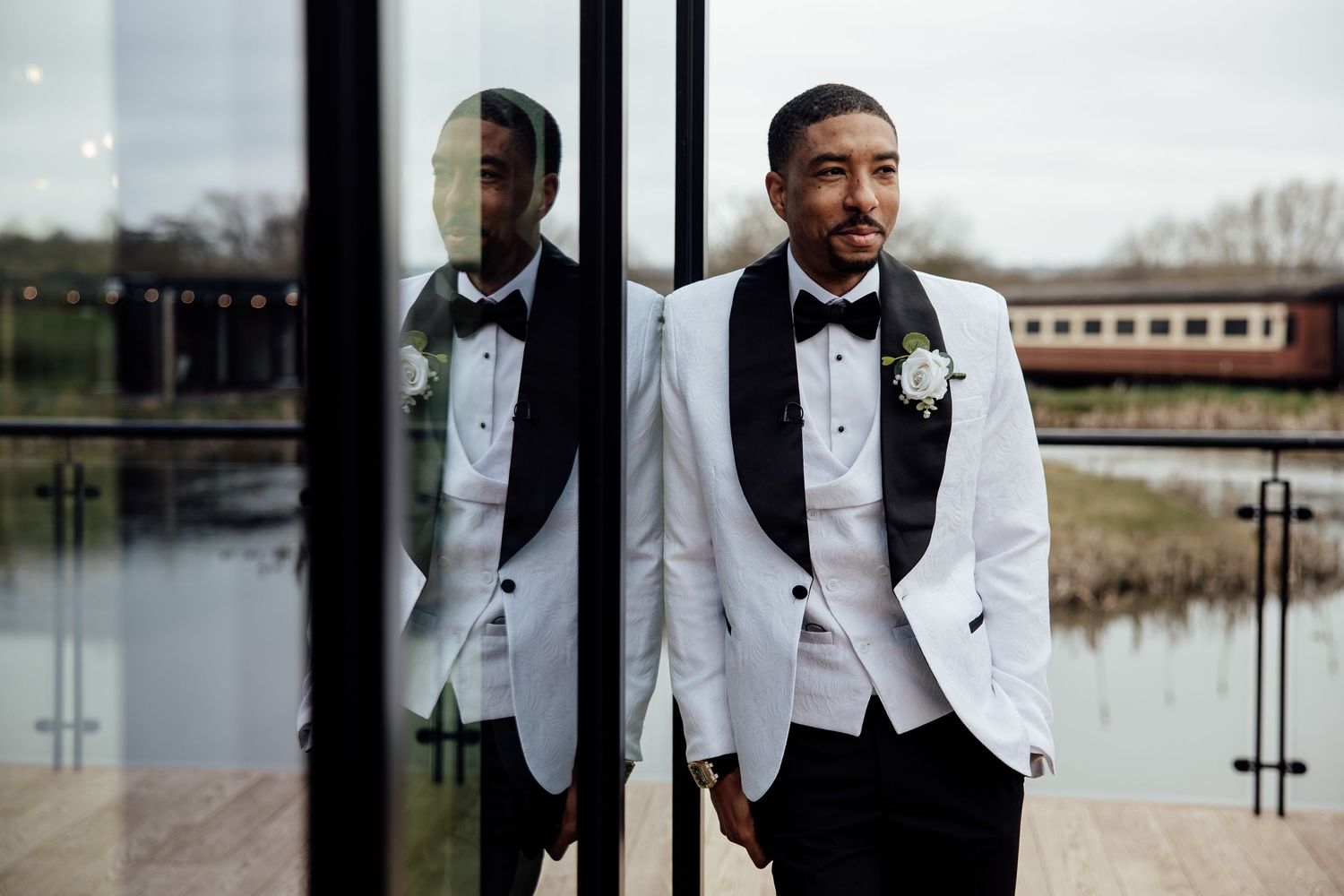 Groom in white tuxedo with black bow tie posing by glass doors with reflection.