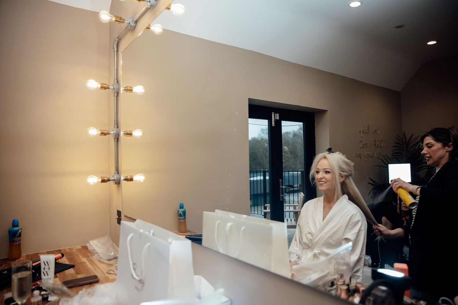 Bride in white robe getting hair styled at vanity mirror with Hollywood lights before wedding.