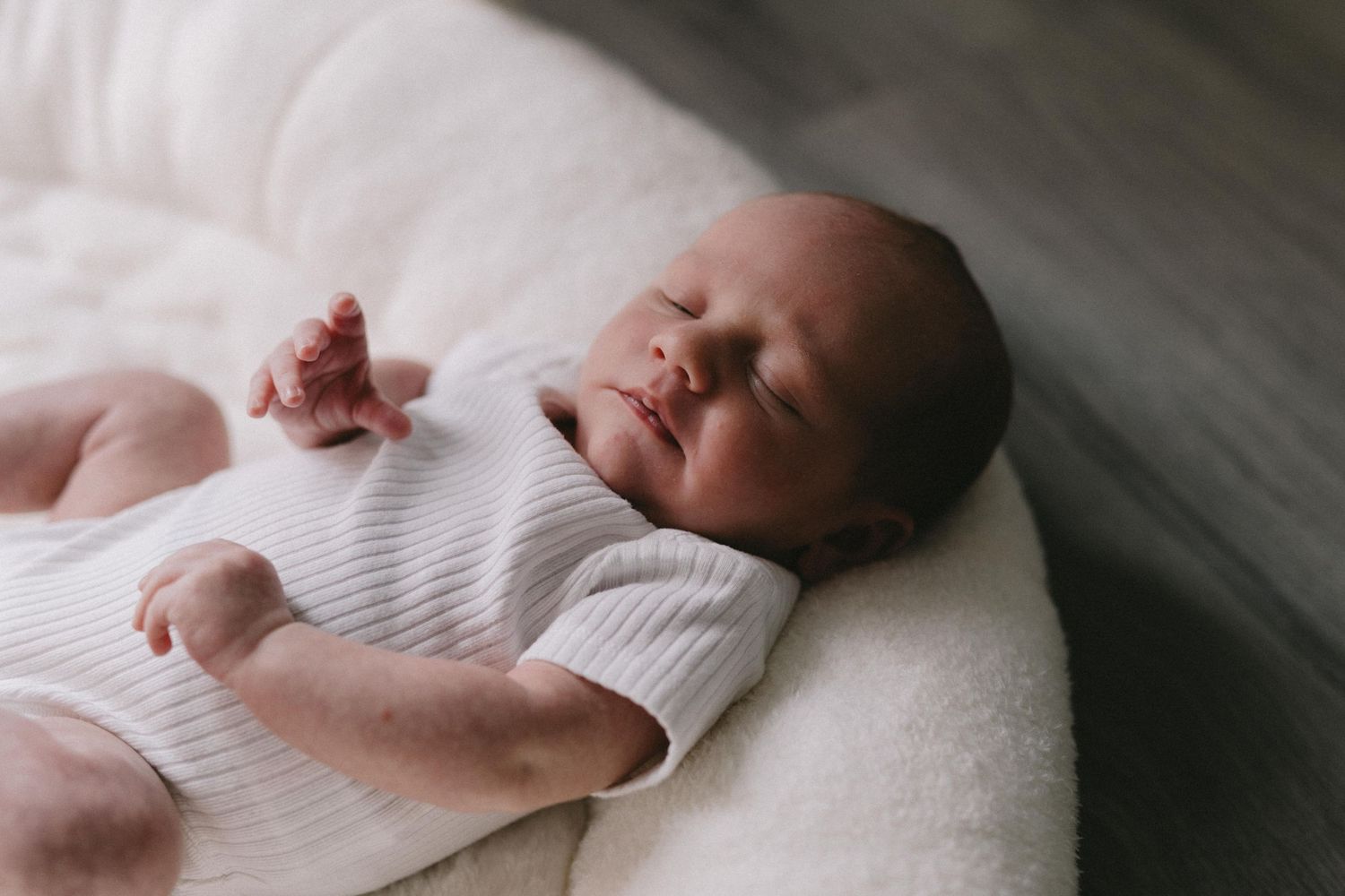 A peaceful newborn baby sleeping in a white nest, wearing a striped onesie with one tiny hand raised.