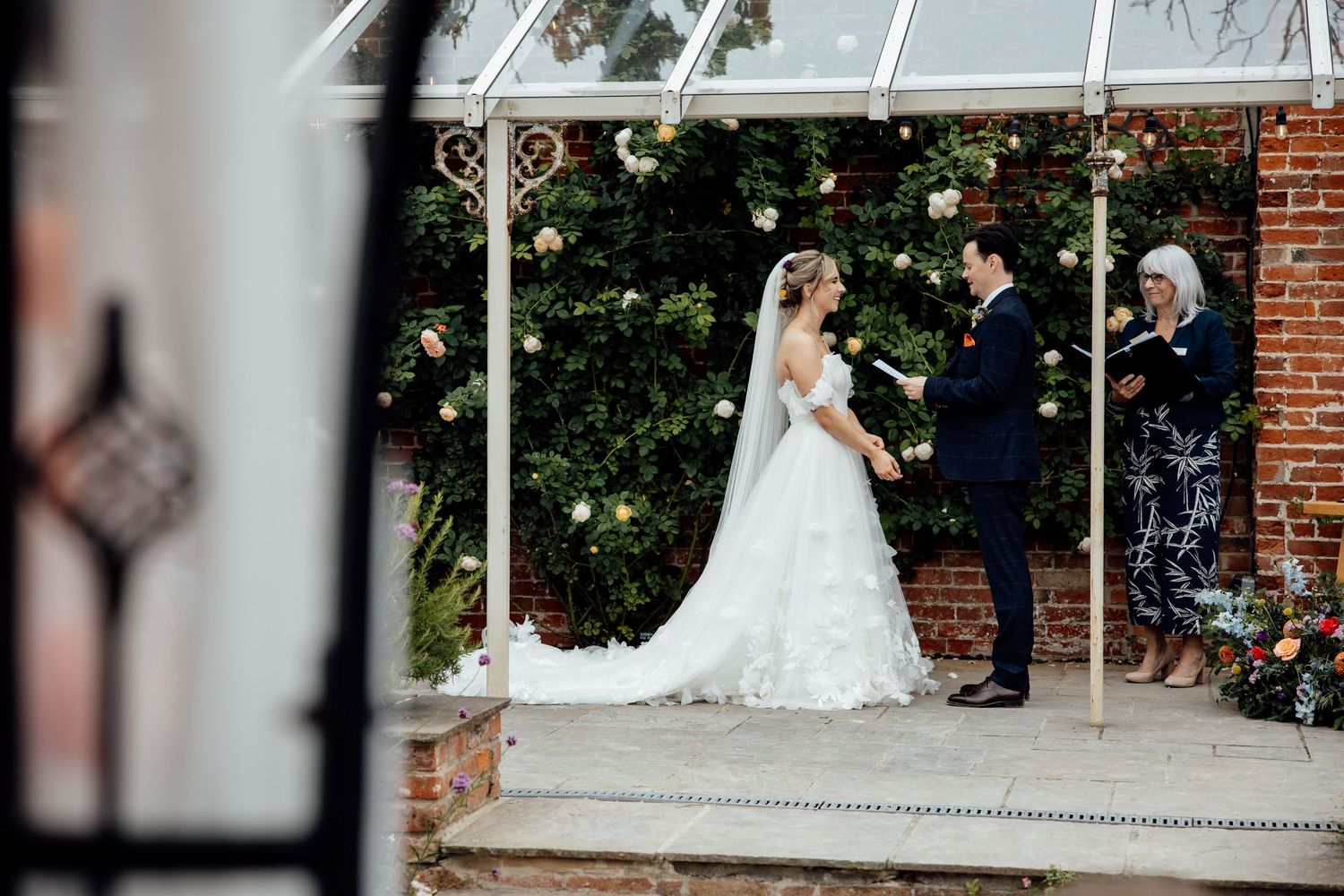 A wedding ceremony taking place under a glass conservatory with brick walls and greenery.