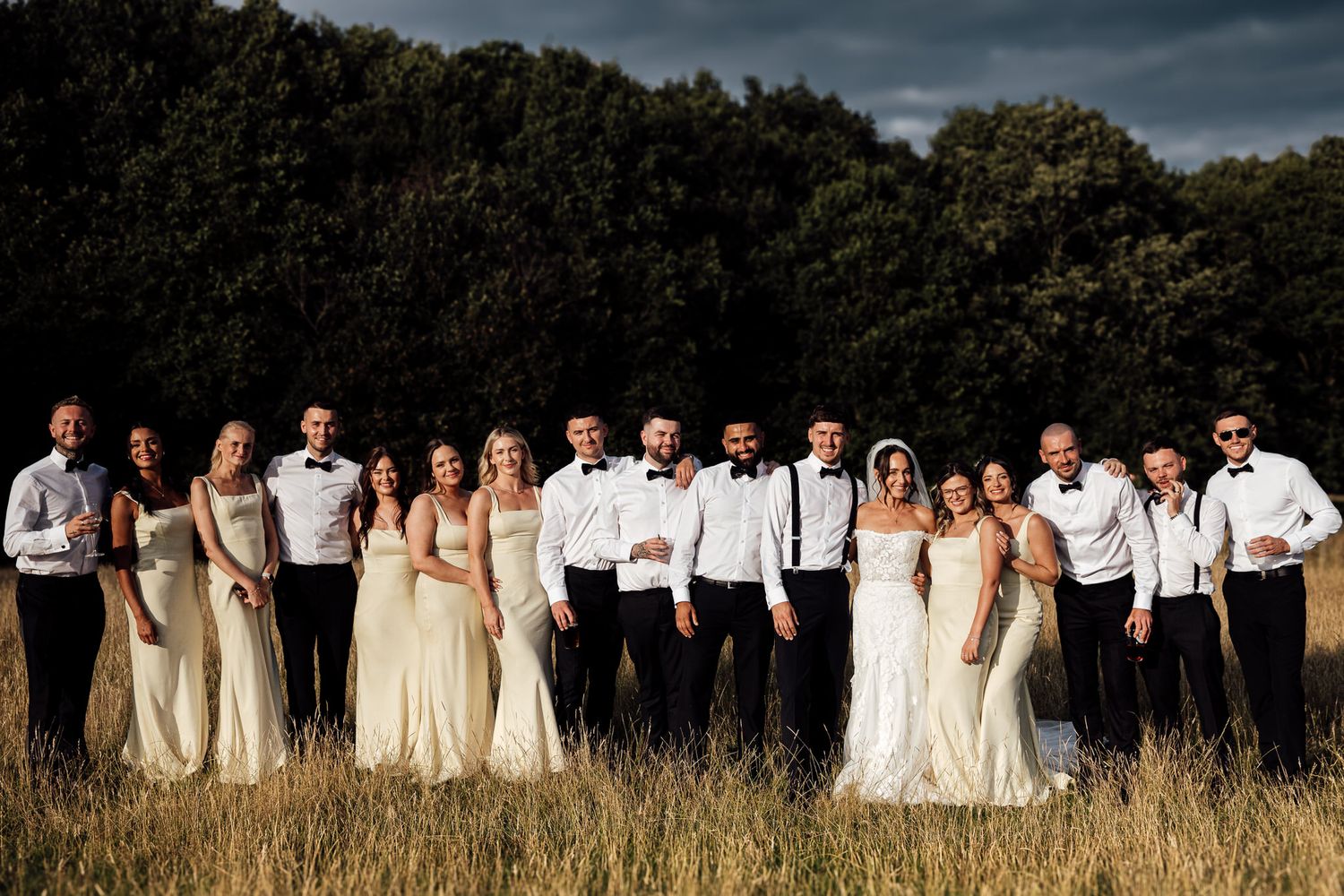 A wedding party in white dresses and dark suits poses together in a field at sunset.