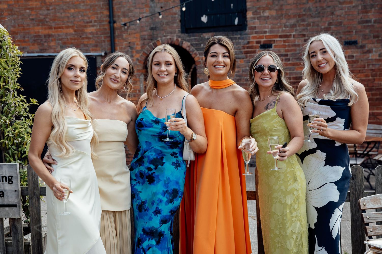 Bridesmaids wearing colorful dresses in orange, blue and yellow pose together against brick wall.