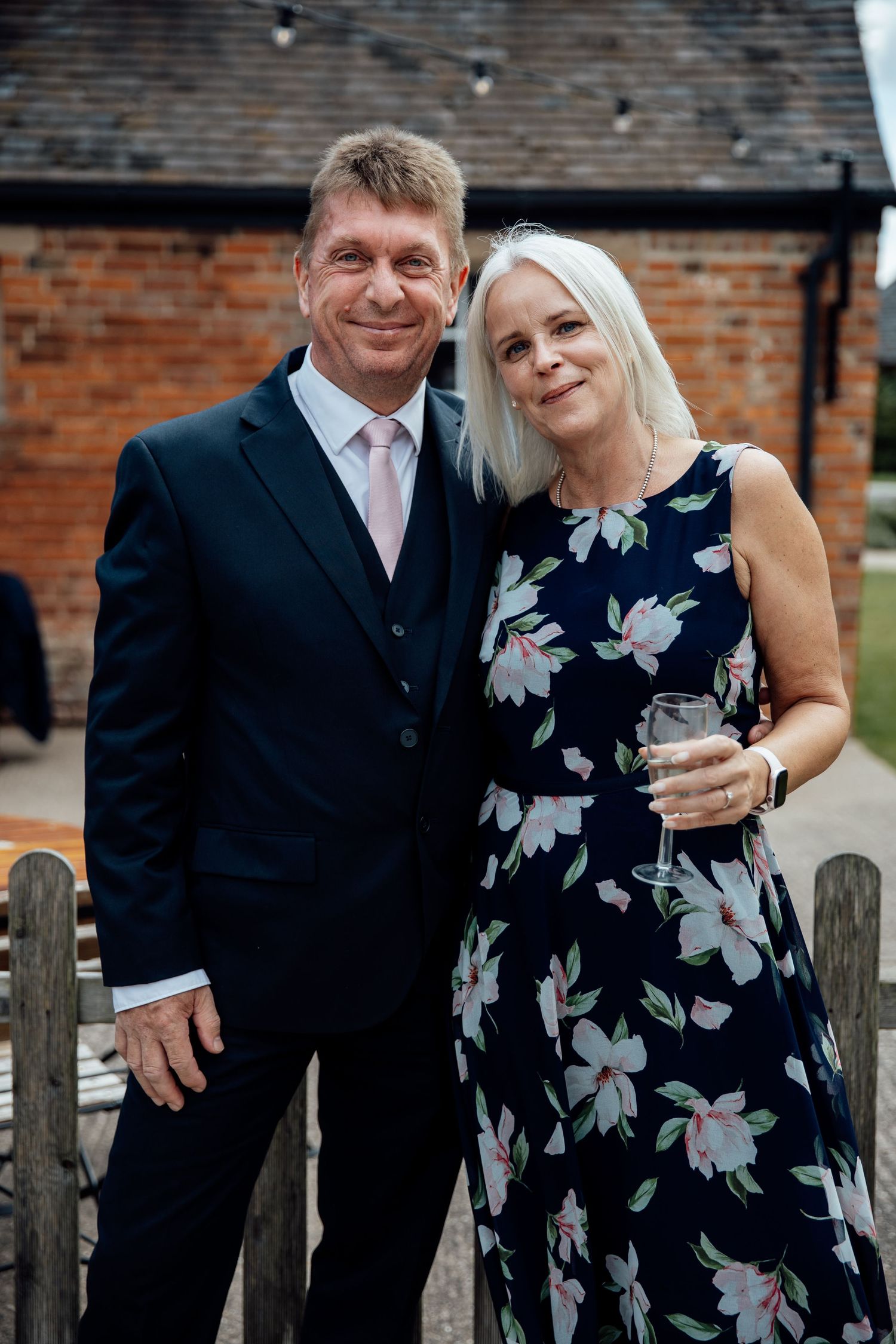 Couple in formal attire shares intimate moment against brick wall backdrop.