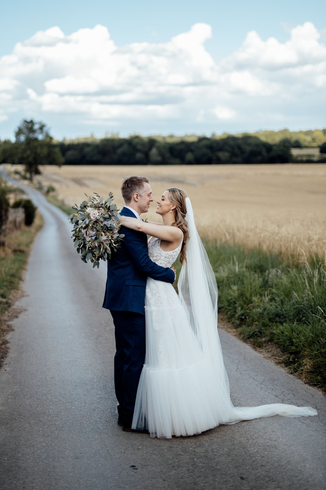 Newlyweds share a romantic moment on a rural road surrounded by wheat fields.