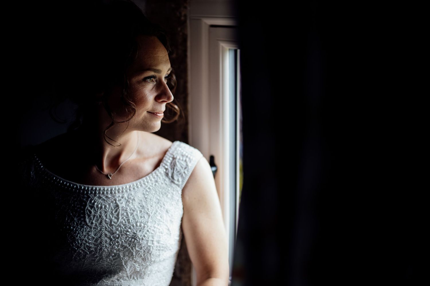 A contemplative figure in a white lace dress gazes out of a window in dramatic lighting.