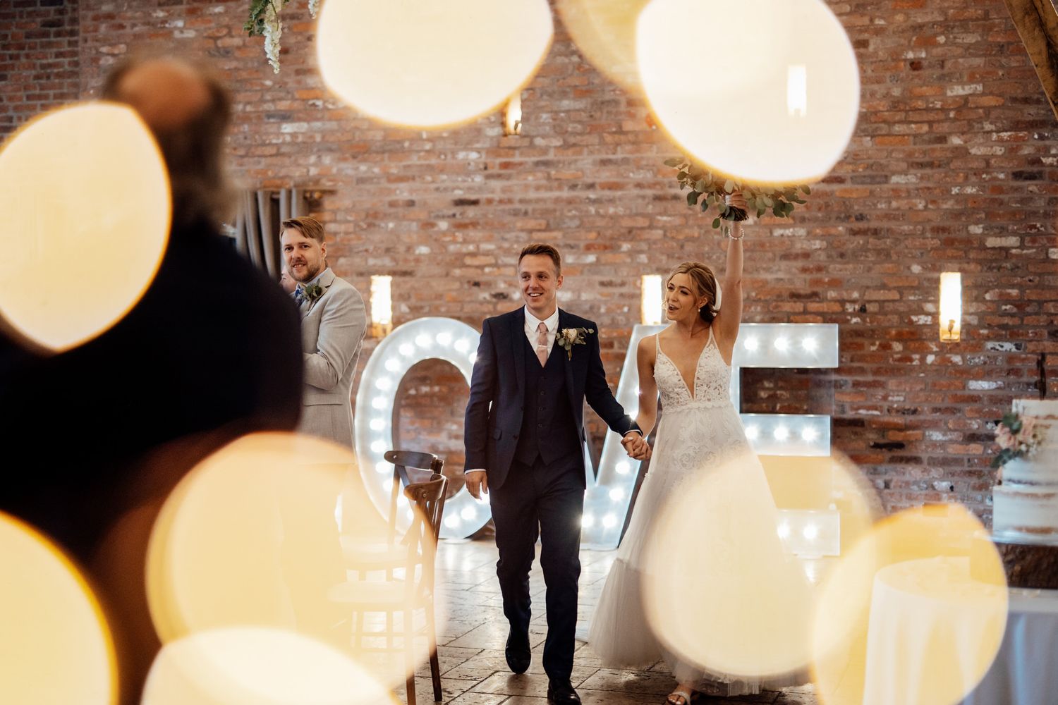 Wedding celebration in brick venue with bokeh lights and illuminated LOVE sign.