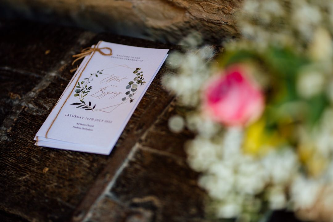 Wedding ceremony program rests on rustic stone surface with blurred floral decorations in background.