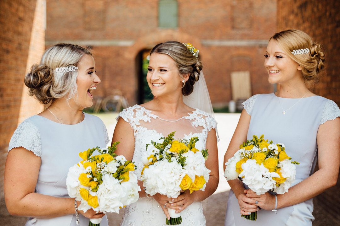 Three women hold beautiful yellow and white wedding bouquets against a brick wall backdrop.