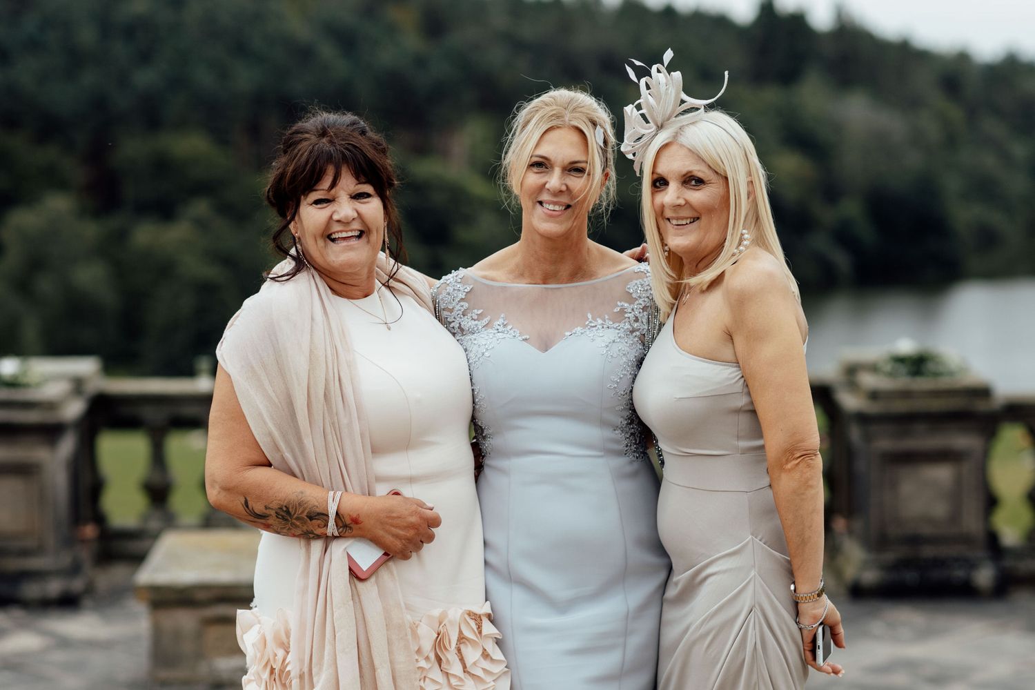 Bridesmaids in pastel dresses pose together by lakeside on wedding day.