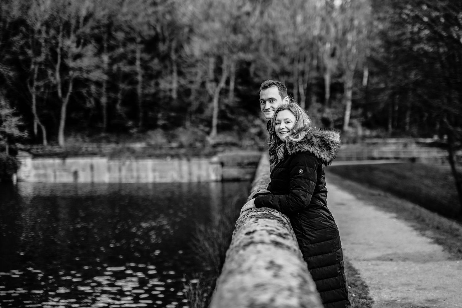 Black and white photo of couple walking along waterfront path surrounded by bare trees.