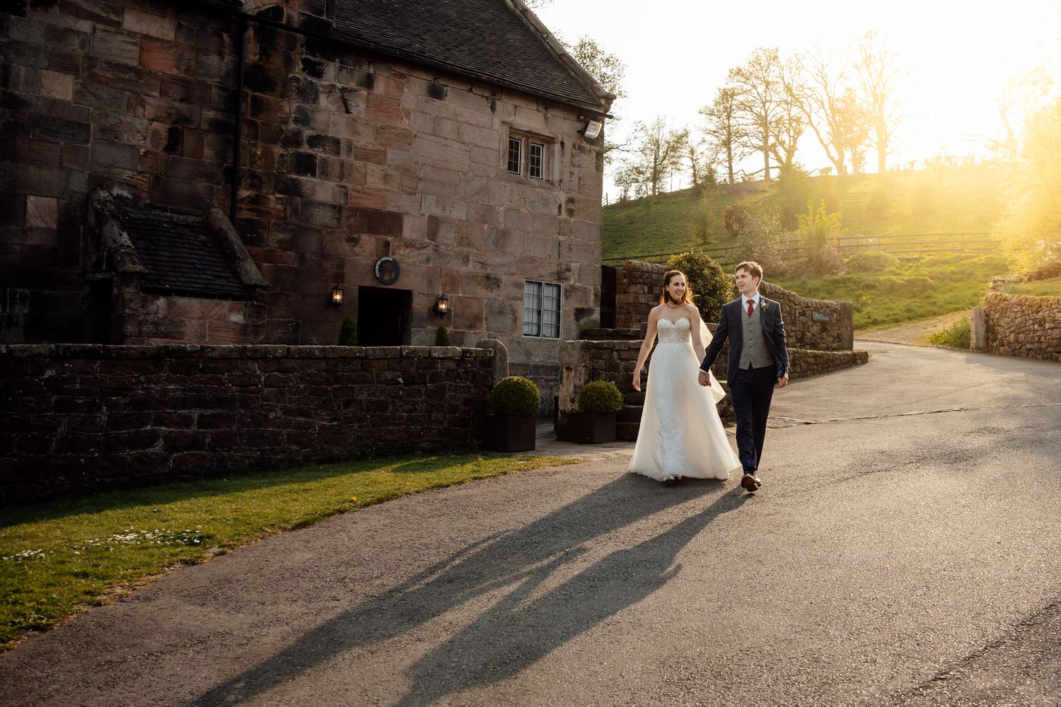 Couple walking along stone path at sunset near historic building during wedding photoshoot.