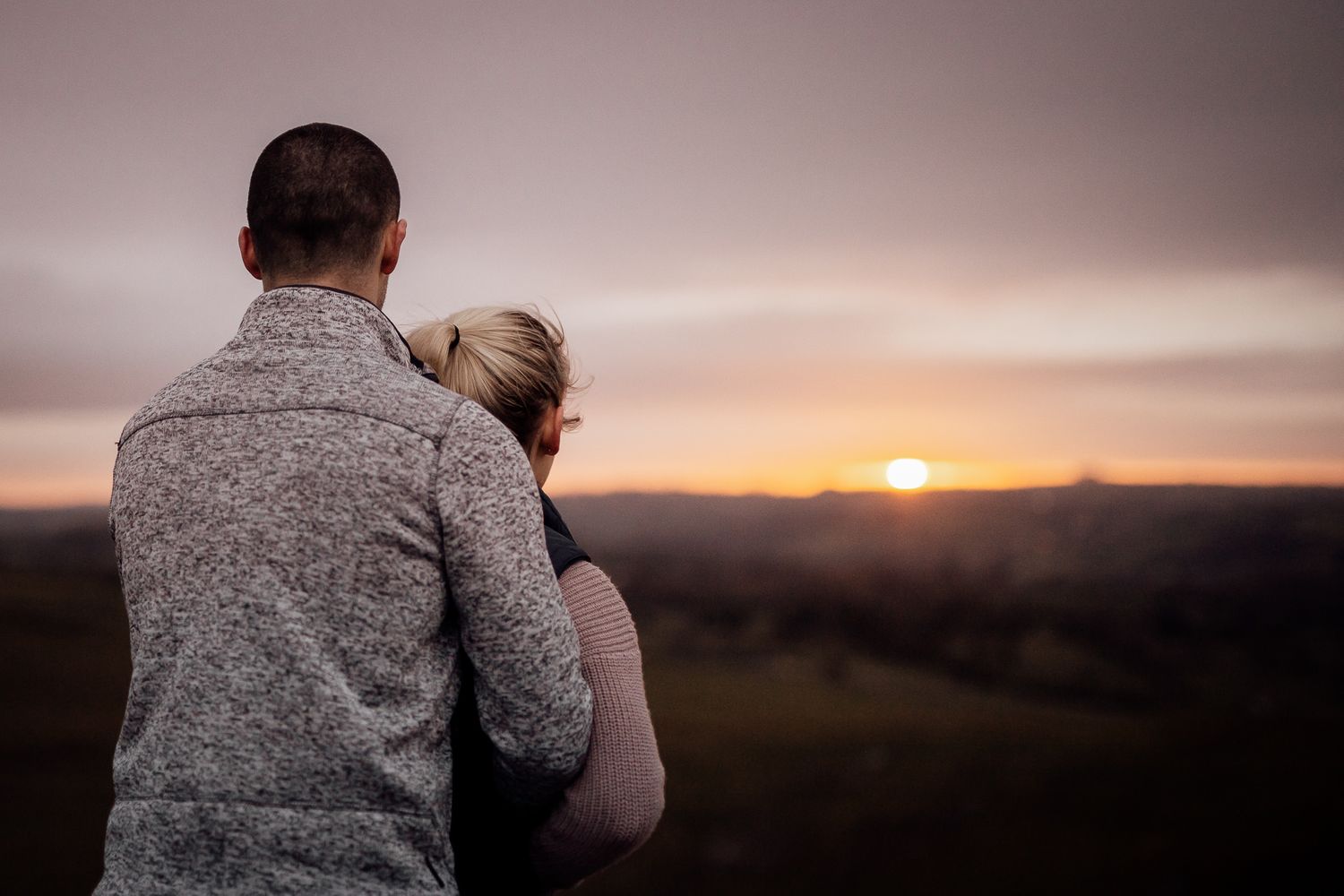 A couple embraces while watching a golden sunset over rolling hills.