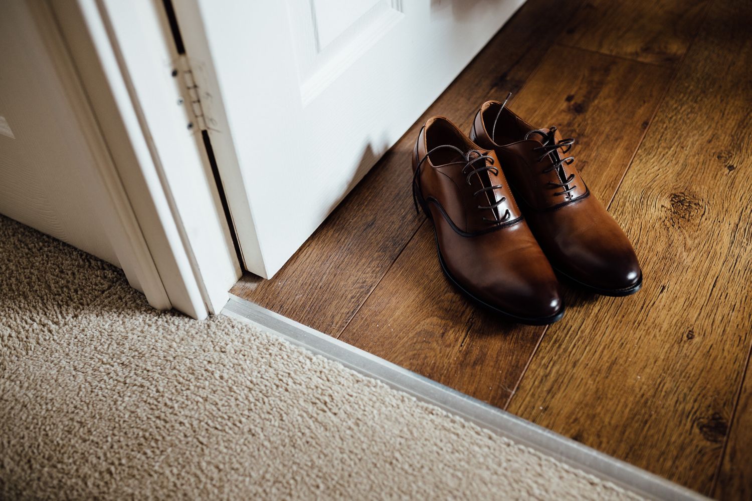 Brown leather dress shoes placed on wooden floor near white door.