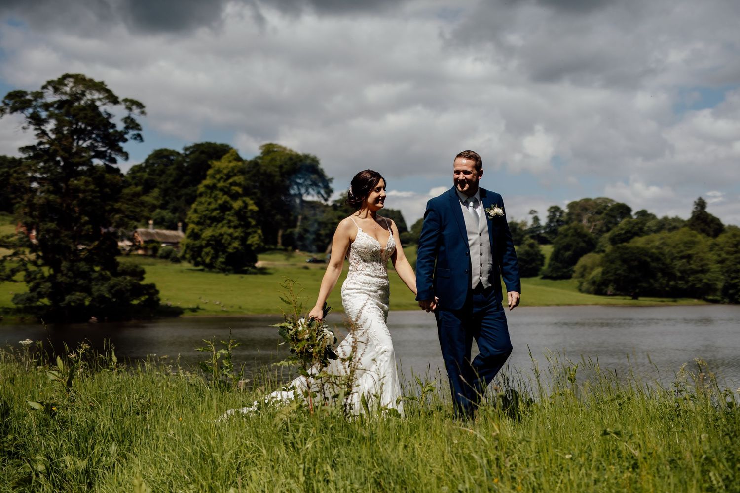Newlyweds walking together through a scenic meadow with a lake and cloudy sky in the background.