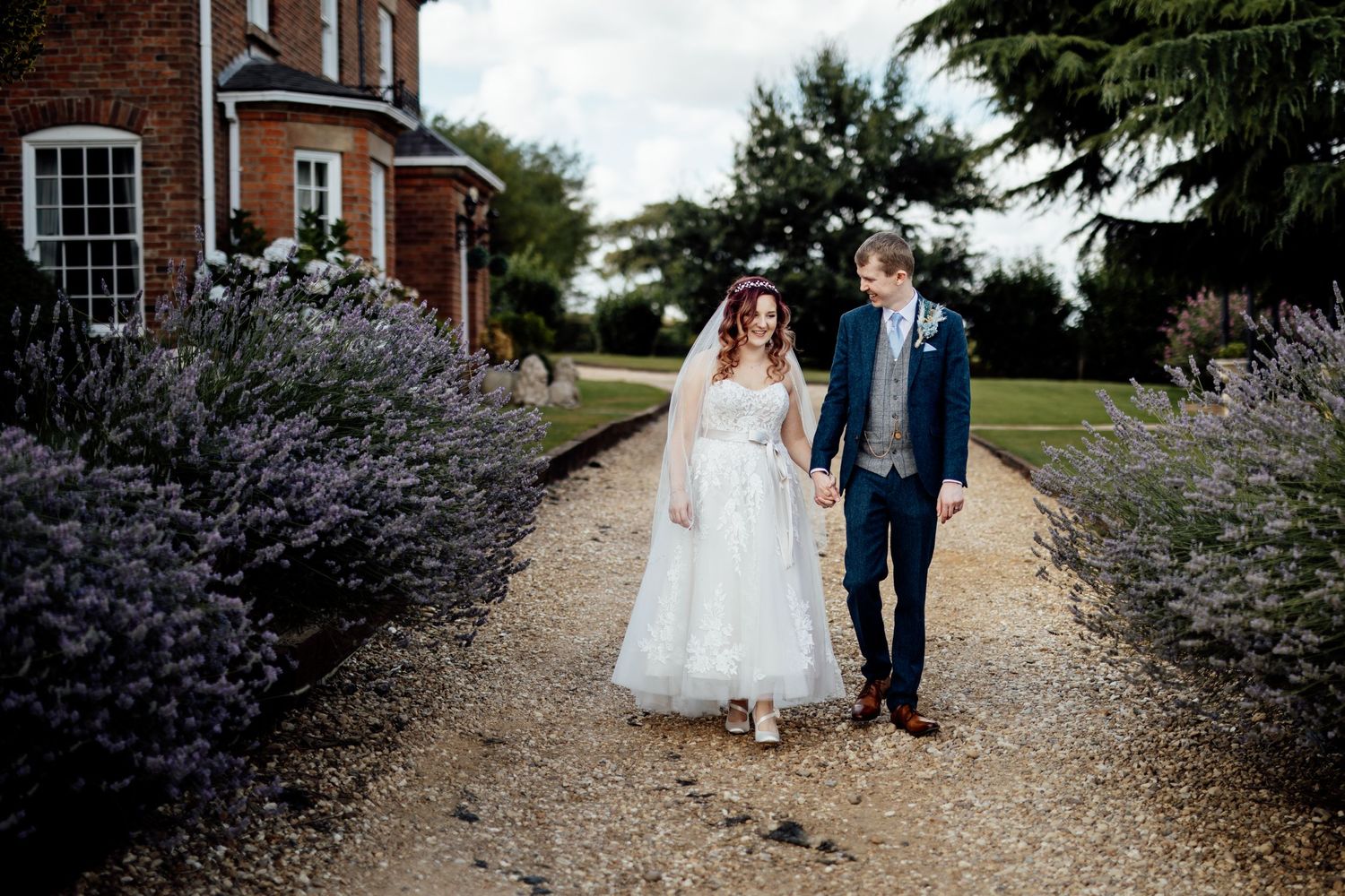 Couple walking down lavender-lined gravel path near brick building.