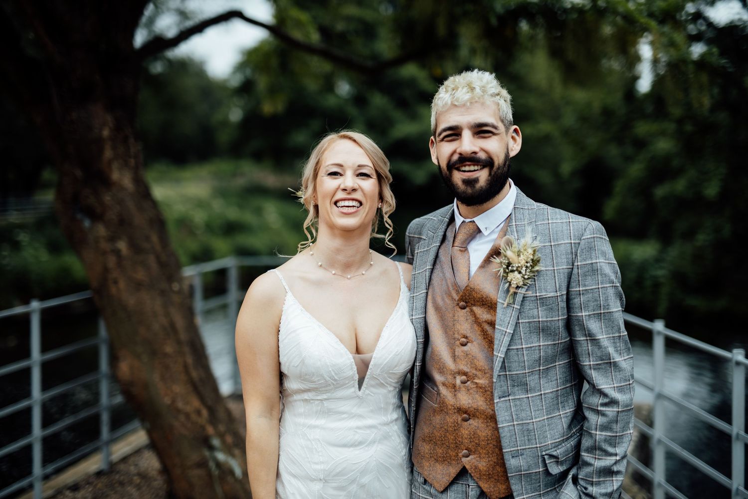 Wedding party poses outdoors on metal walkway surrounded by green trees.