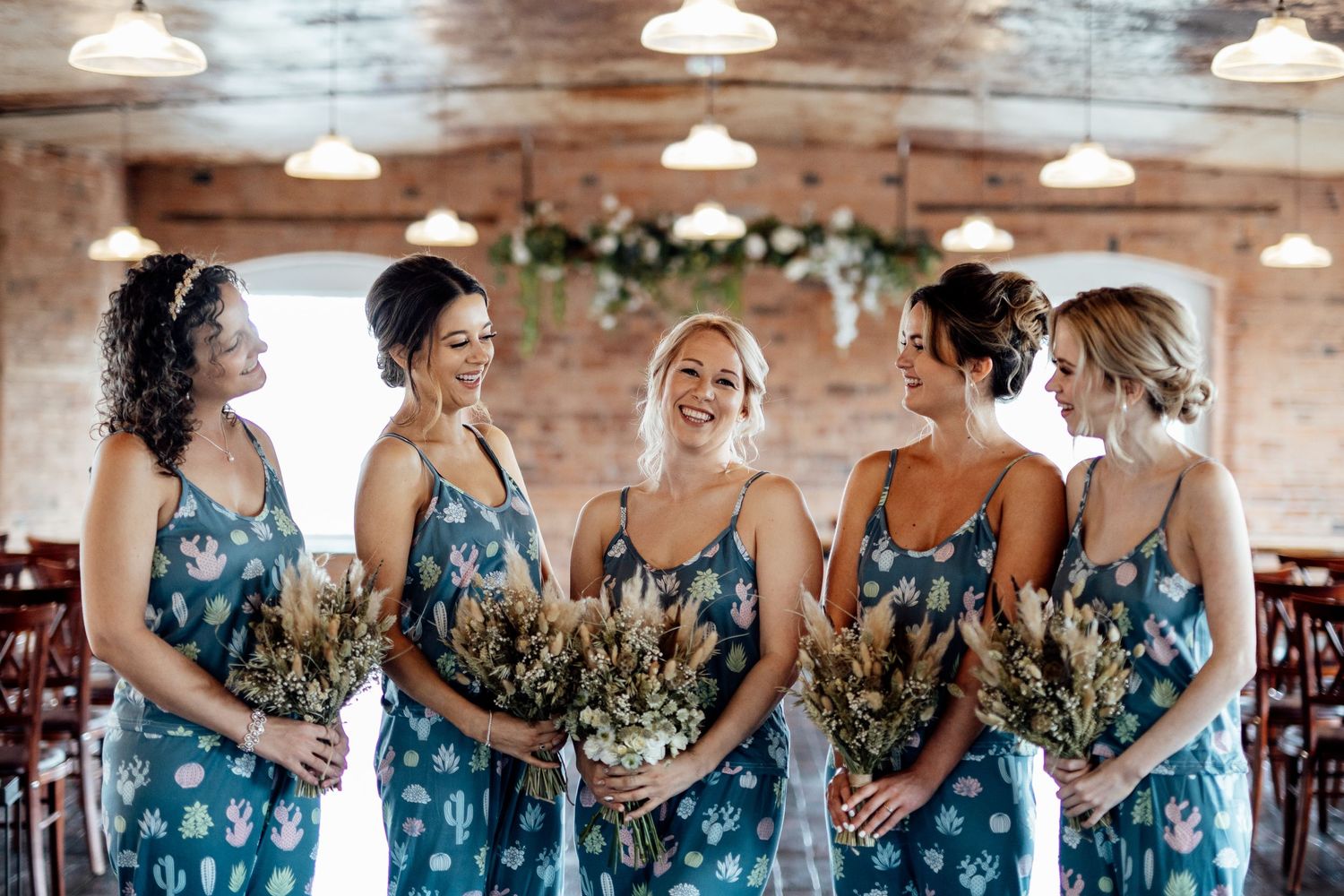 Group wearing matching blue floral dresses holding bouquets in a rustic brick venue.
