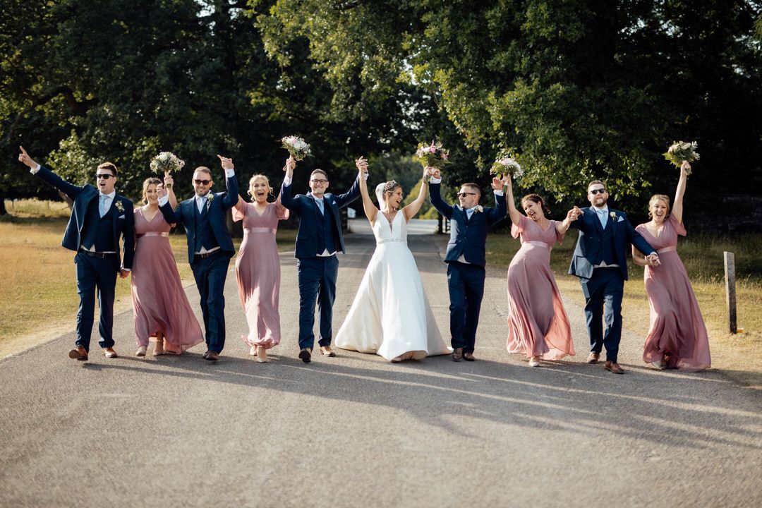 Wedding party jumps in celebration on path wearing navy suits and dusty rose dresses.