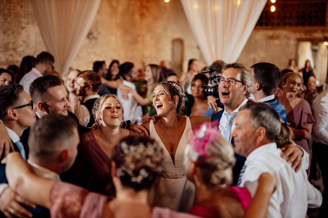 Guests enjoy dancing together at a wedding reception in a rustic brick venue with white draping.