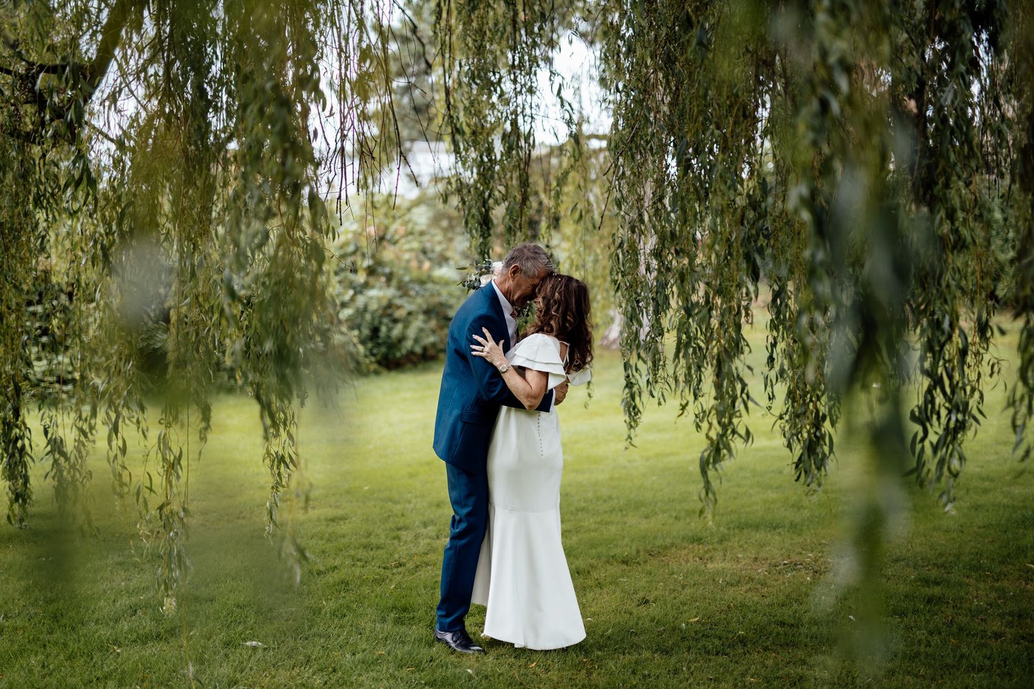 Romantic couple embraces under cascading willow tree branches in a lush garden setting.