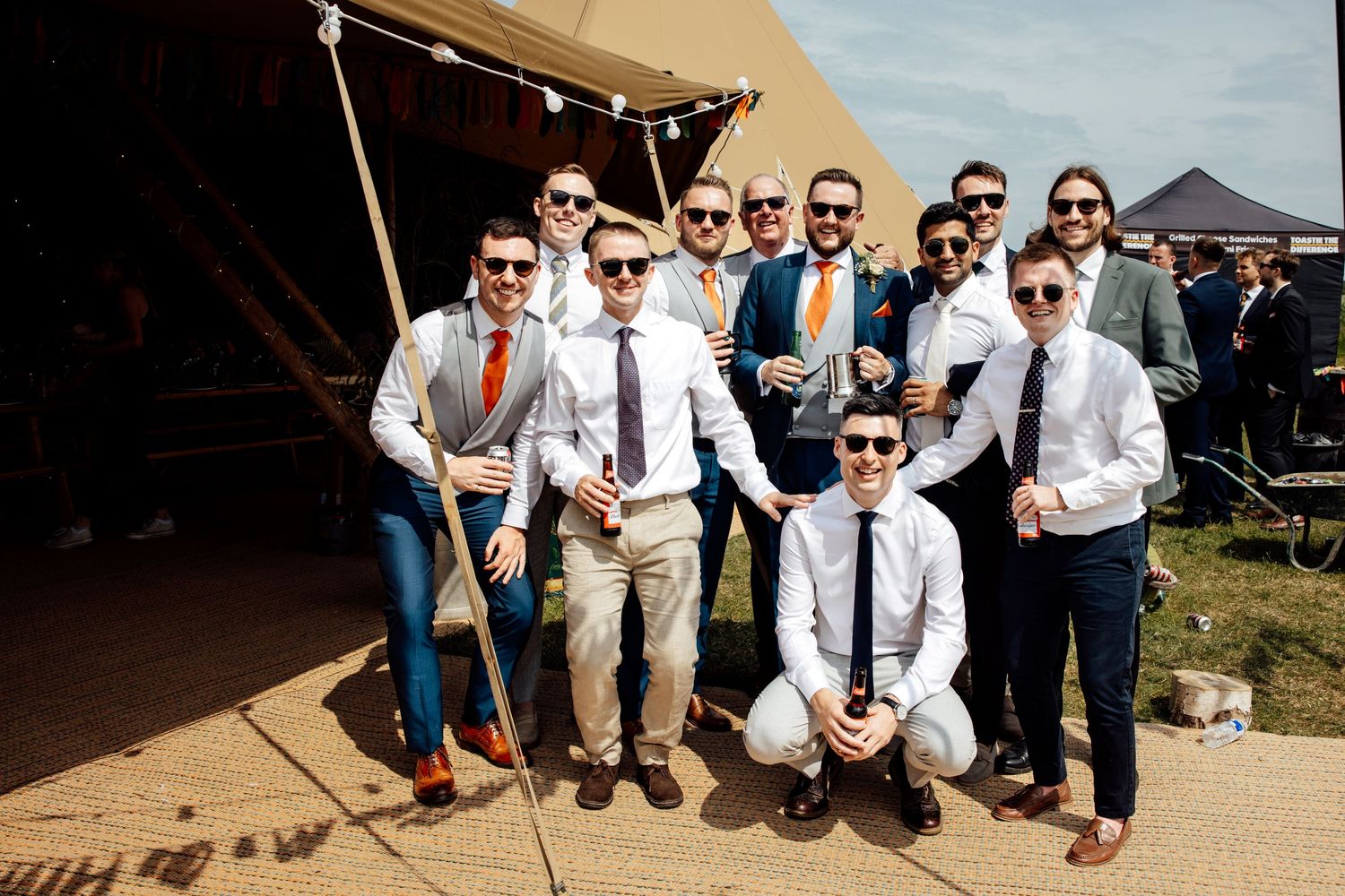 Group photo of wedding party members in sunglasses and formal attire outside tent.