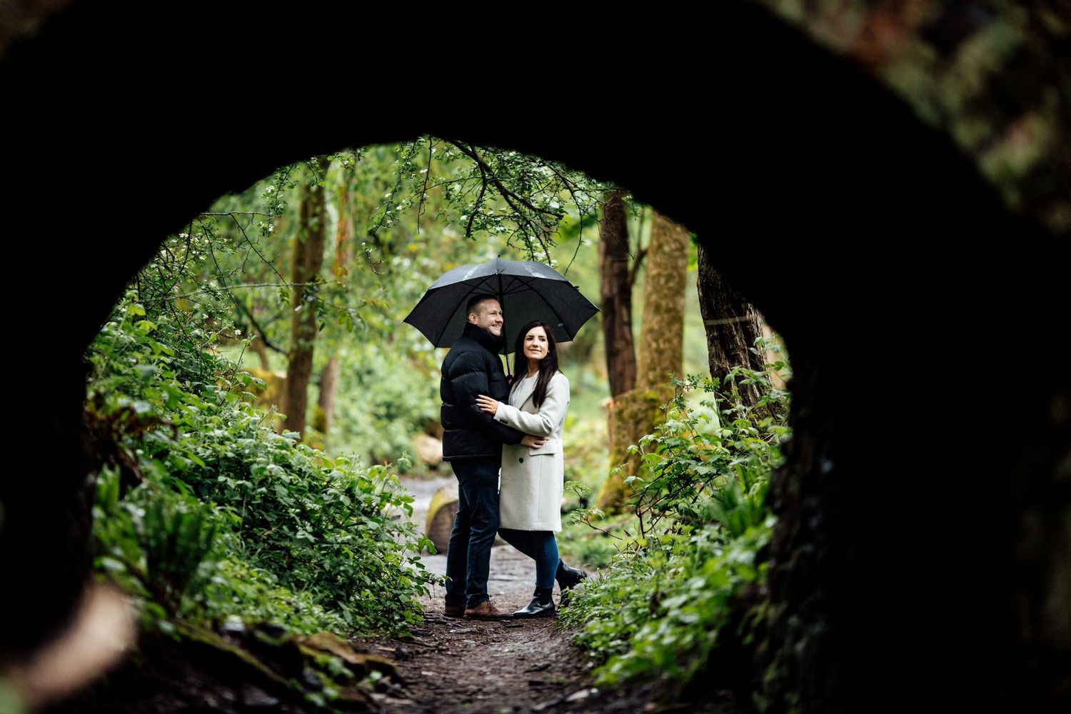 A couple stands together in a dark tunnel framed by natural greenery.