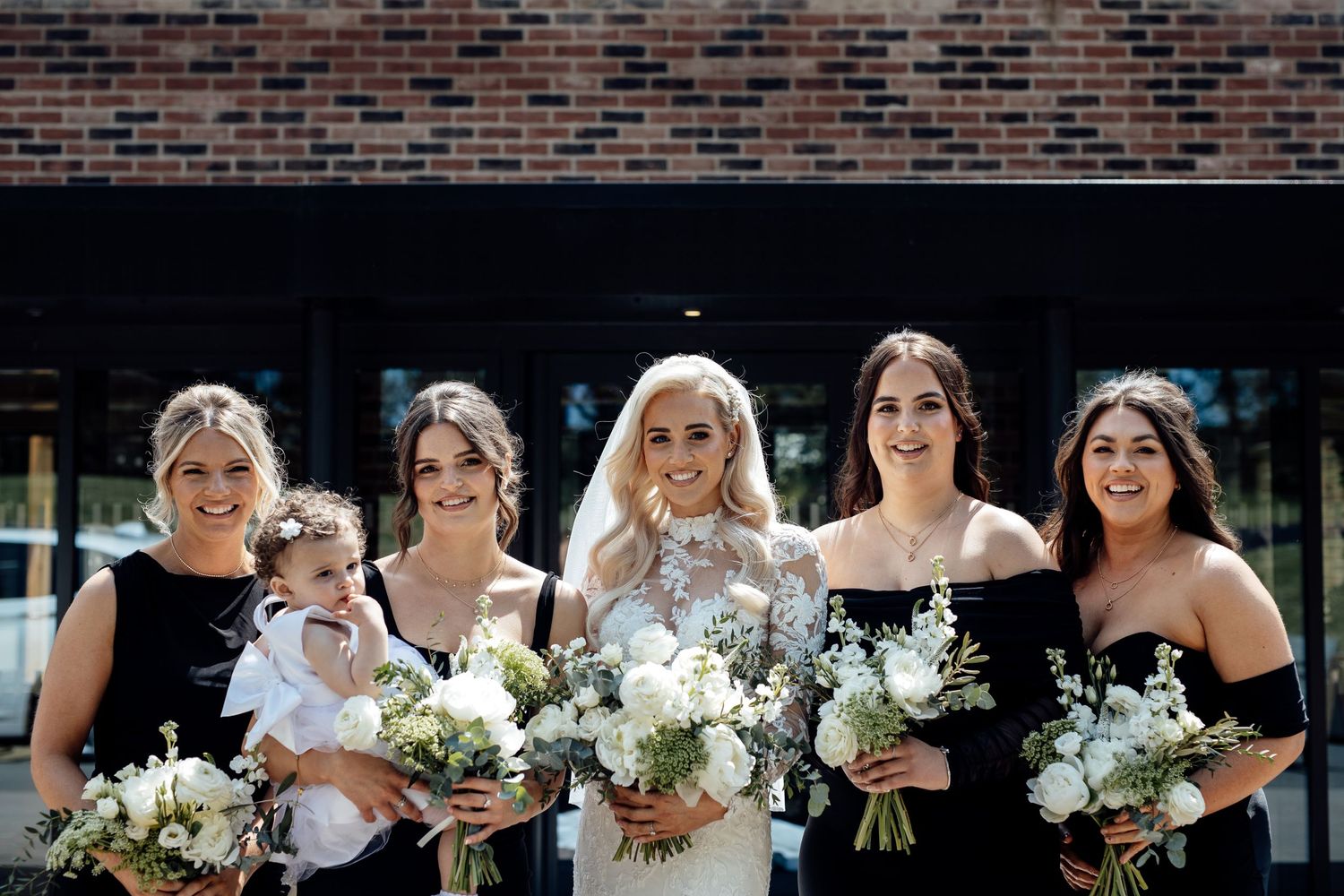 Bridesmaids in black dresses pose together holding white floral bouquets against a brick wall.