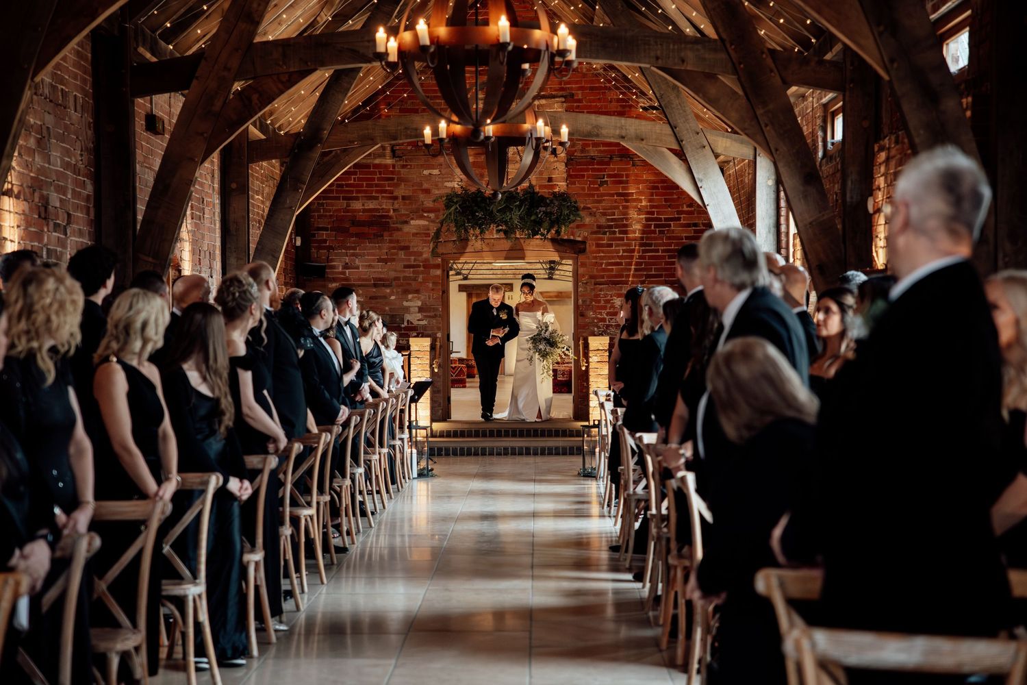 Long aisle lined with wooden chairs in a rustic brick venue illuminated by chandeliers.