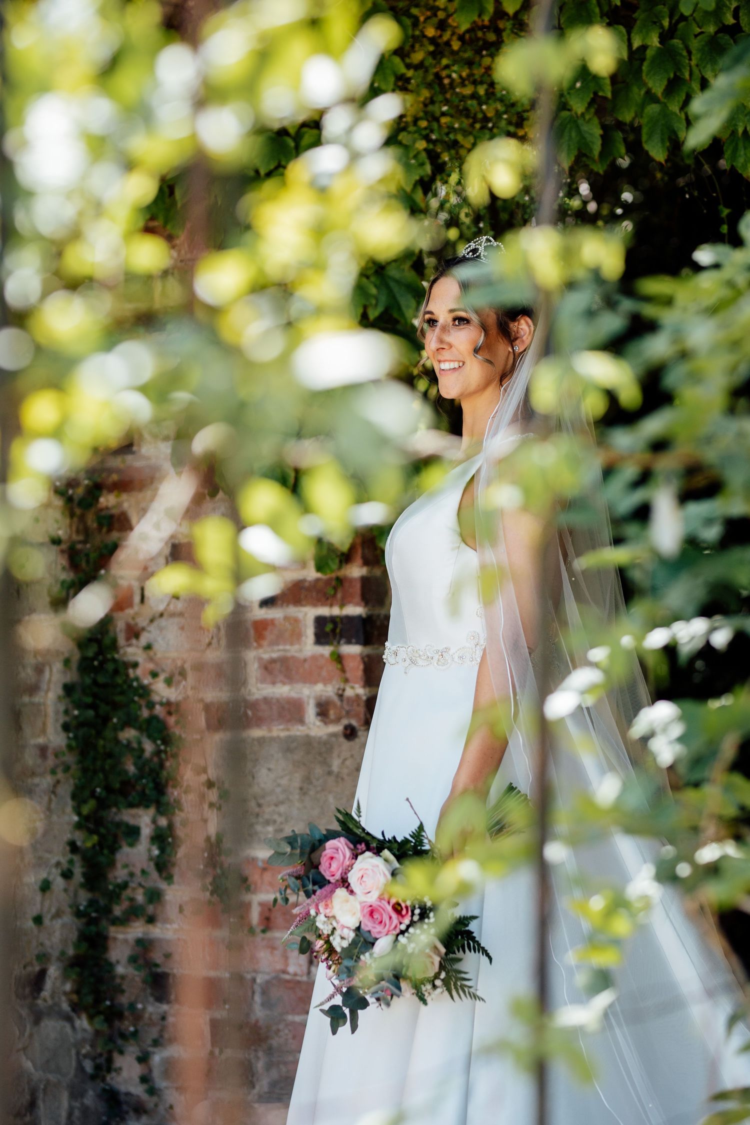 A bride in a white dress holds a pink and white bouquet against a brick wall backdrop with soft bokeh lighting.