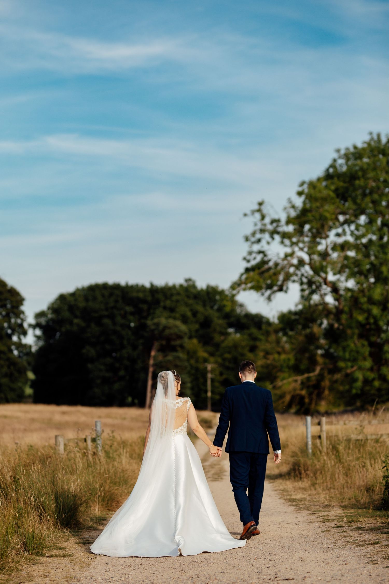 A bride and groom walk hand in hand down a countryside path surrounded by golden fields and trees on their wedding day.
