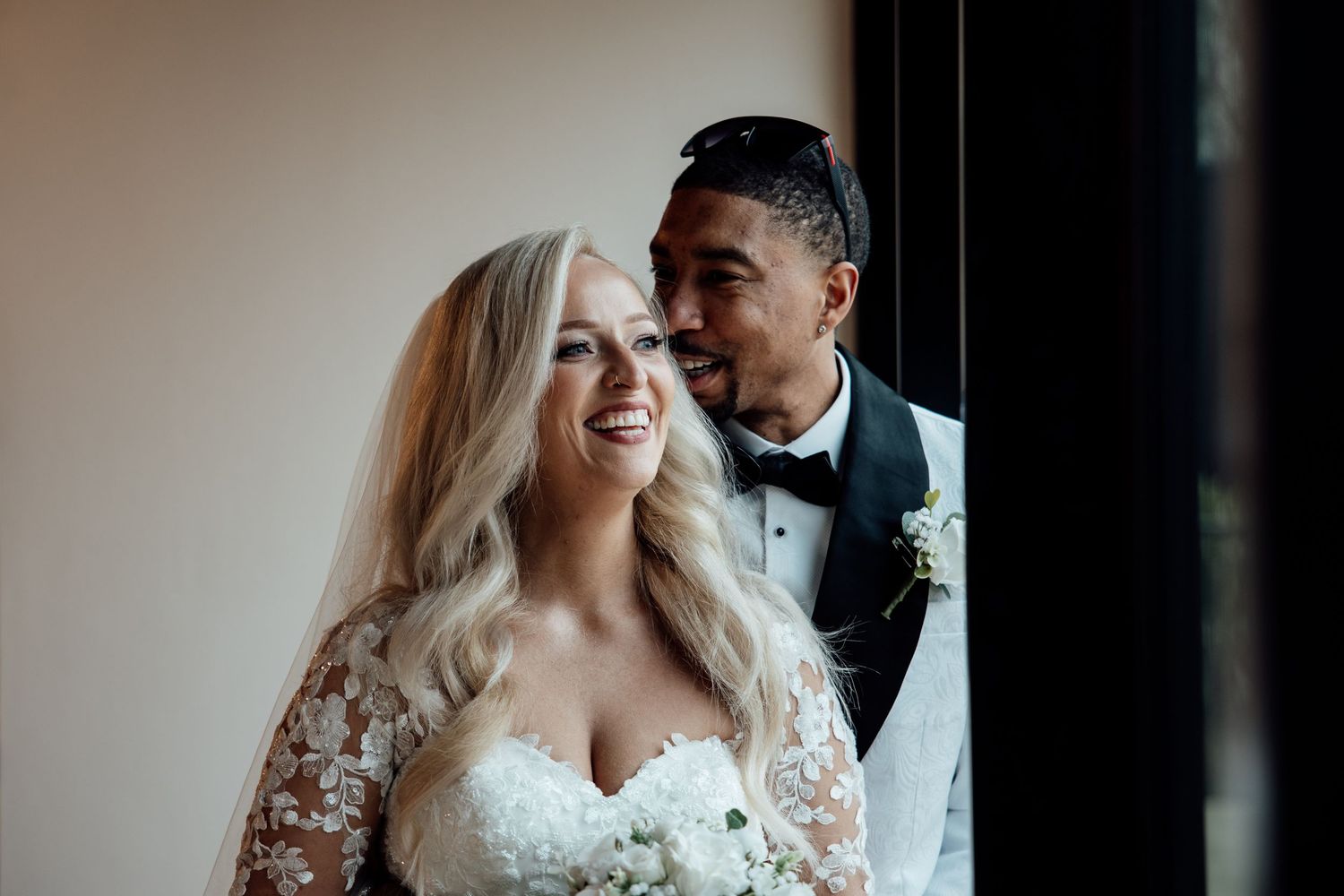 Smiling bride in lace wedding gown with groom in white tuxedo sharing a romantic moment by a window.