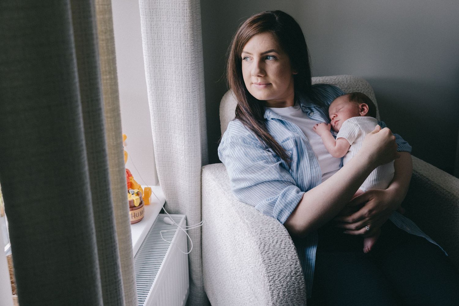 Young mother sitting in armchair holding sleeping newborn baby near window in soft natural light.