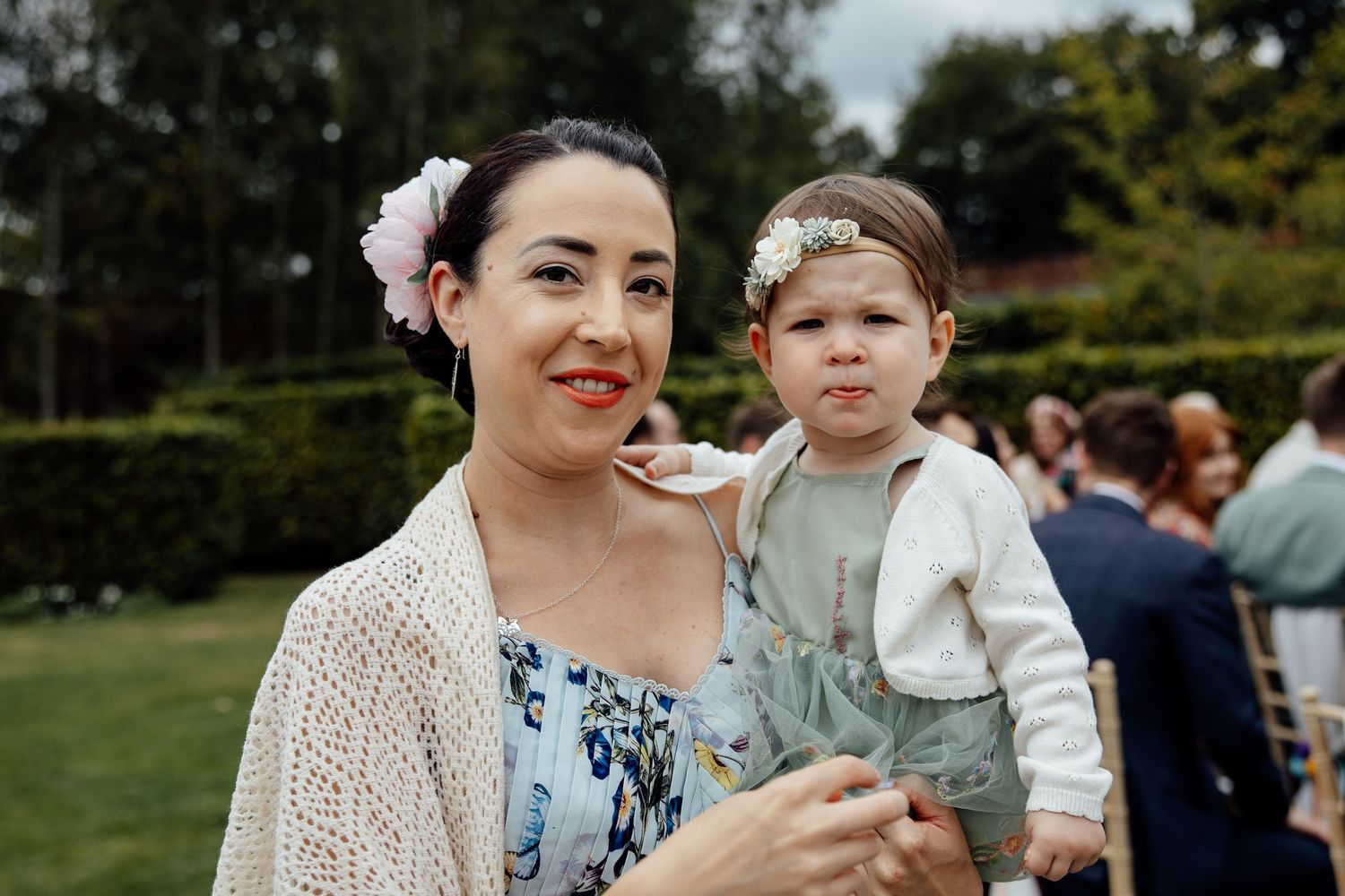 Guest in floral dress holds young child at outdoor garden wedding.