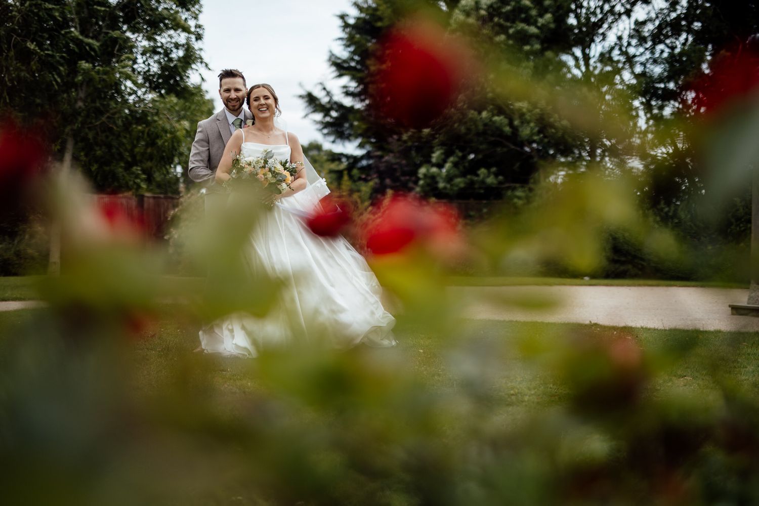 Wedding photography through red flowers with soft focus natural outdoor setting.
