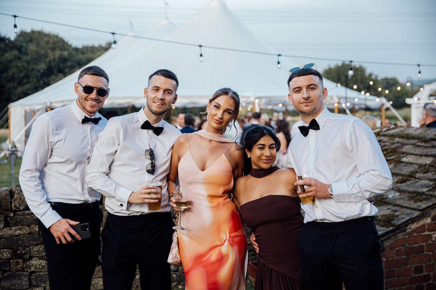 Wedding party poses together outdoors against rustic brick wall with string lights overhead.