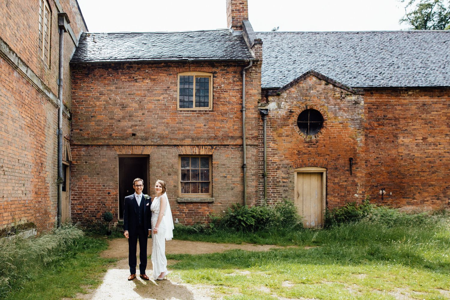 A couple stands in front of a rustic brick building with vintage wooden doors and windows.