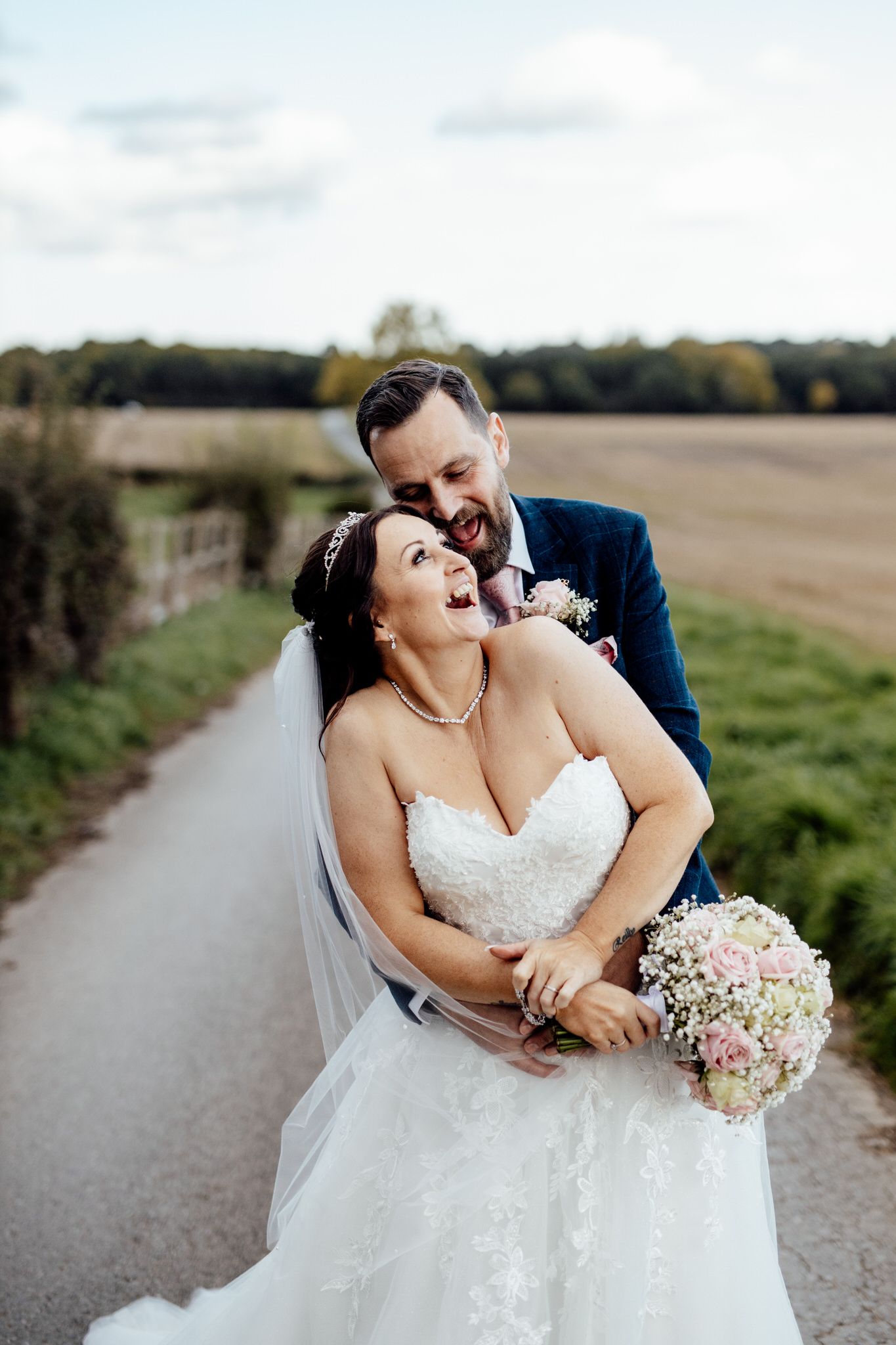 Newlyweds share intimate moment on country path with pink floral bouquet.