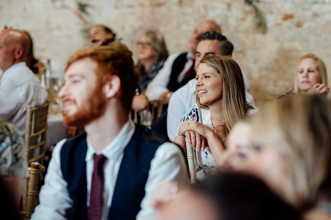 Wedding guests seated at tables listen attentively during reception speeches in rustic brick venue.