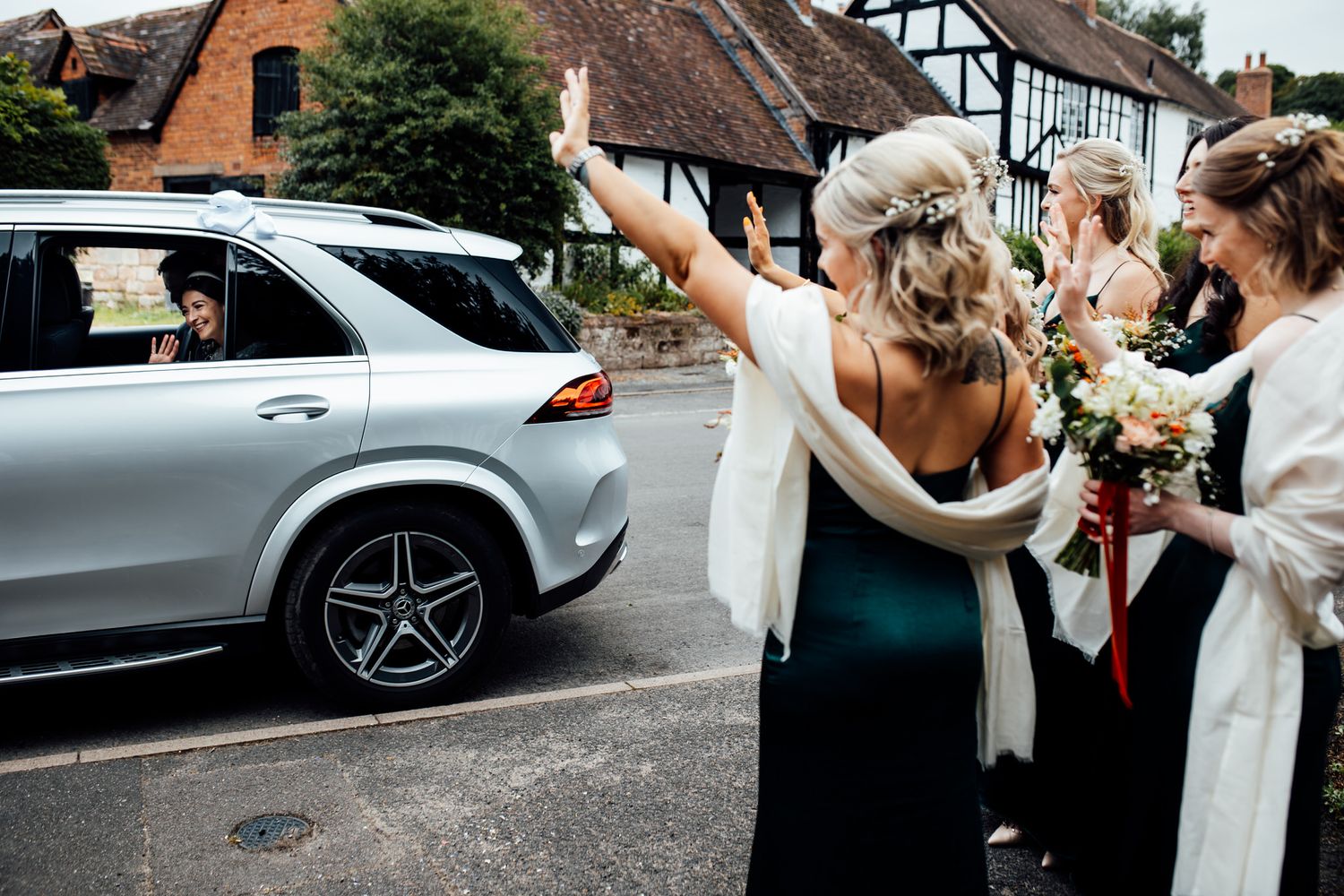 Wedding party exits venue while guests celebrate with confetti near parked cars.
