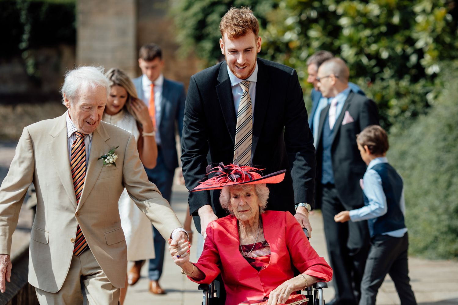 Wedding guests in formal attire gathering outdoors on a sunny day.