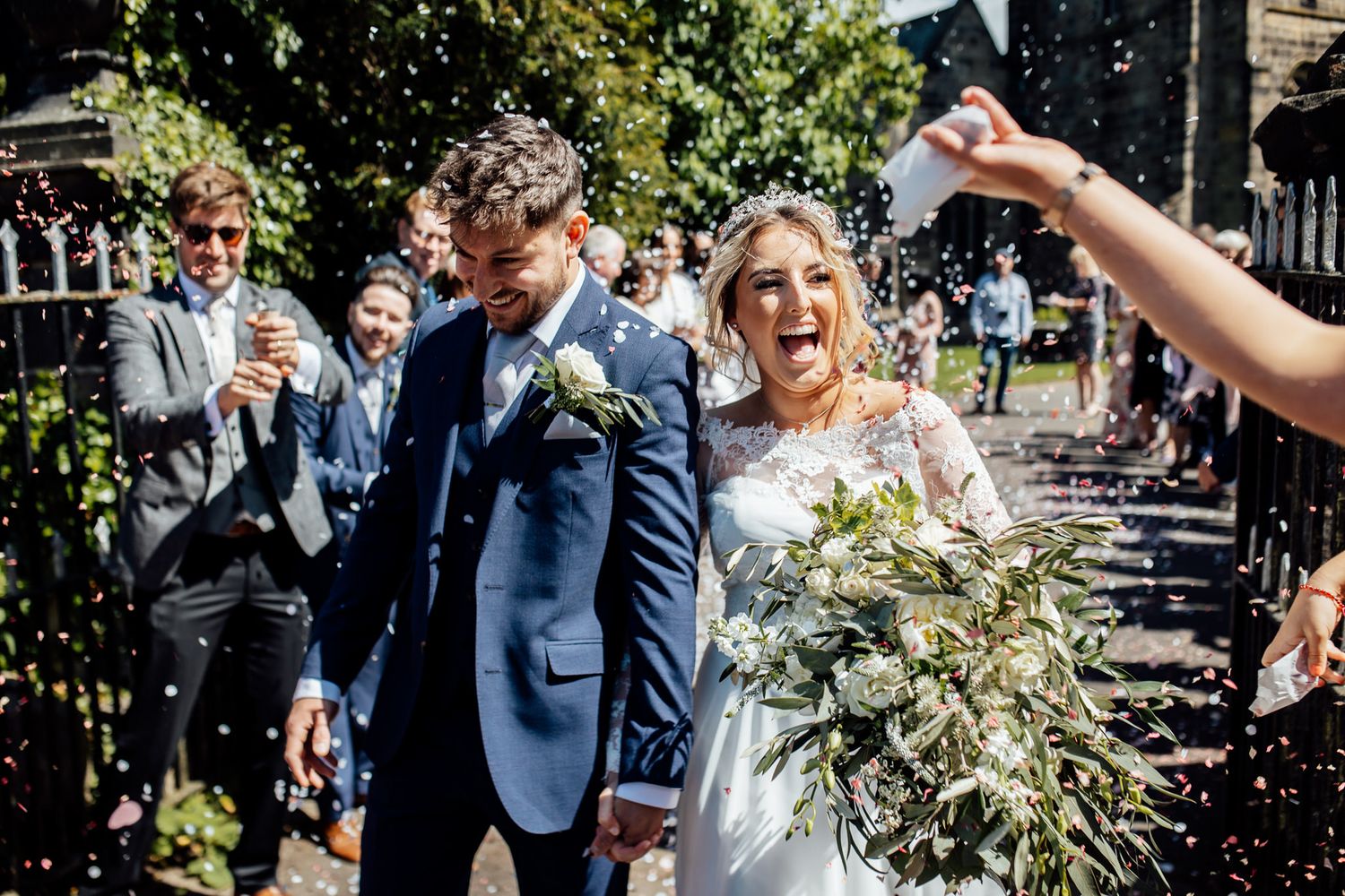 Newlyweds celebrating with guests throwing confetti outside on a sunny day.