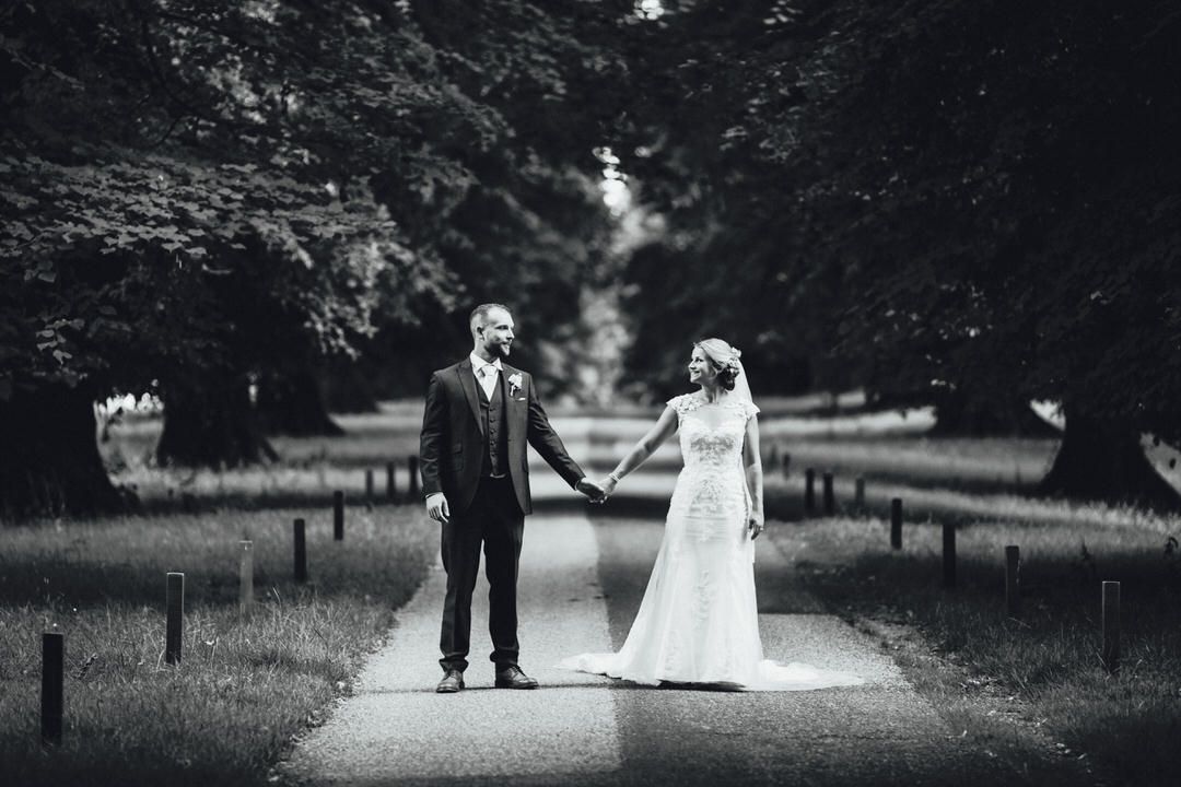 Black and white portrait of newlyweds holding hands on tree-lined path.