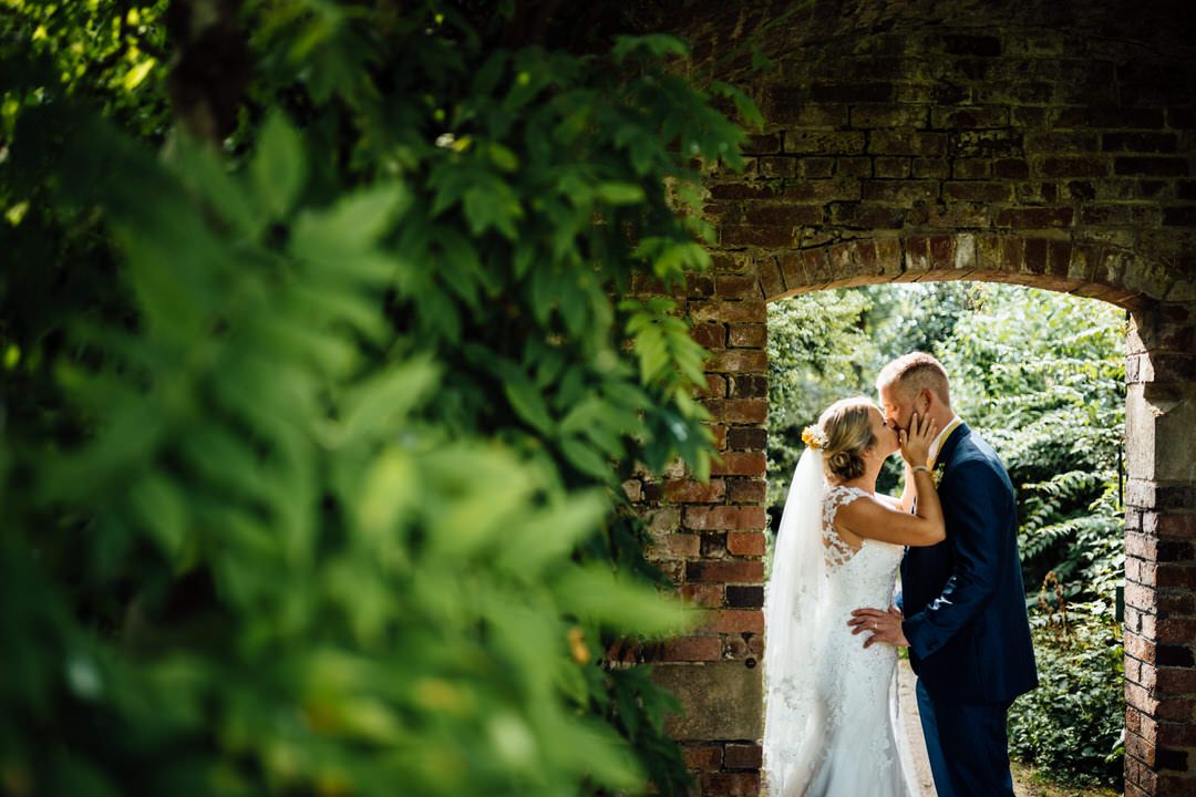 Wedding couple walks through a garden path with brick walls and green foliage.