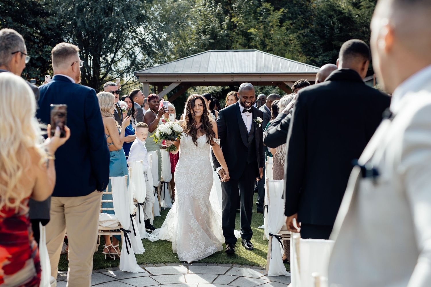 Outdoor wedding ceremony with guests seated in white chairs on a sunny day.