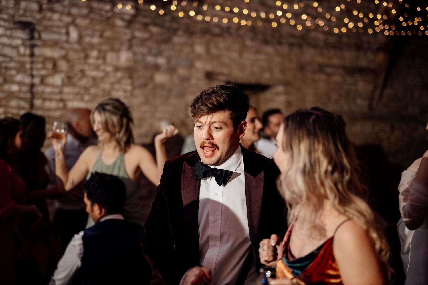 Wedding guest dancing enthusiastically at reception in brick-walled venue with string lights.