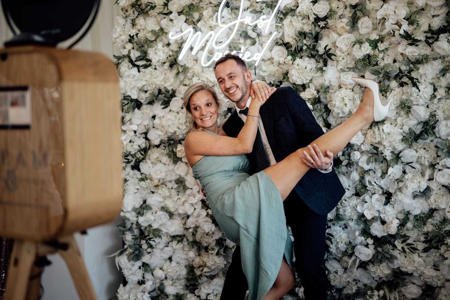 A couple poses playfully in front of a white floral wall backdrop at a wedding celebration.