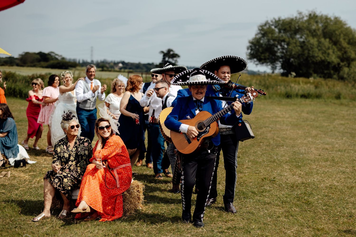 Wedding guests enjoy an outdoor celebration on a grassy field under blue skies.