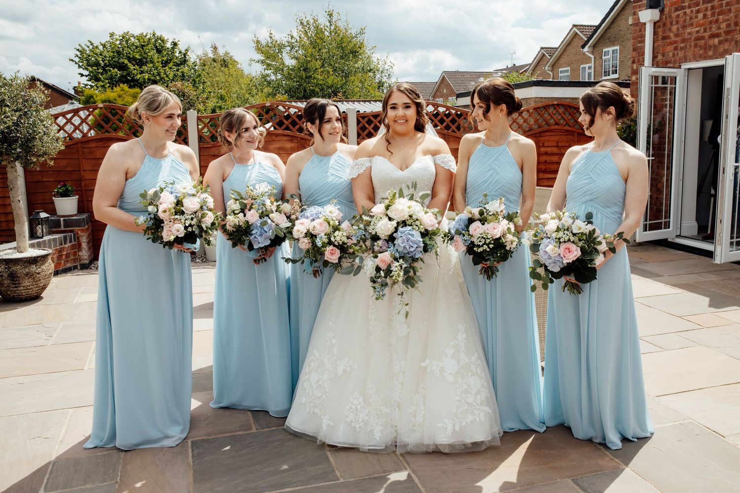 Wedding party in light blue bridesmaid dresses holding white and blue floral bouquets on outdoor terrace.